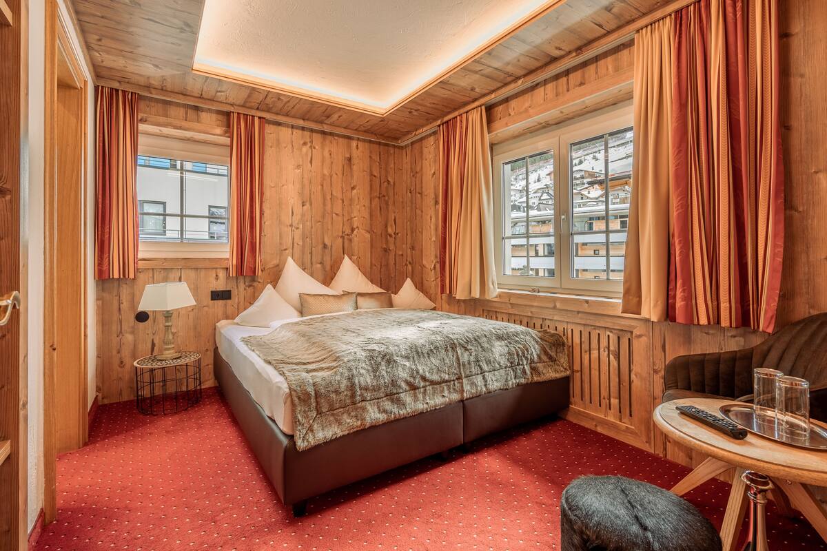 A wooden-paneled room with a bed, a small table with glasses and a remote, and a window with curtains.