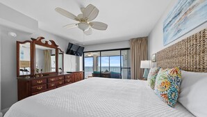 A hotel room with a large bed, a ceiling fan, a TV, a dresser, and a balcony with a view.