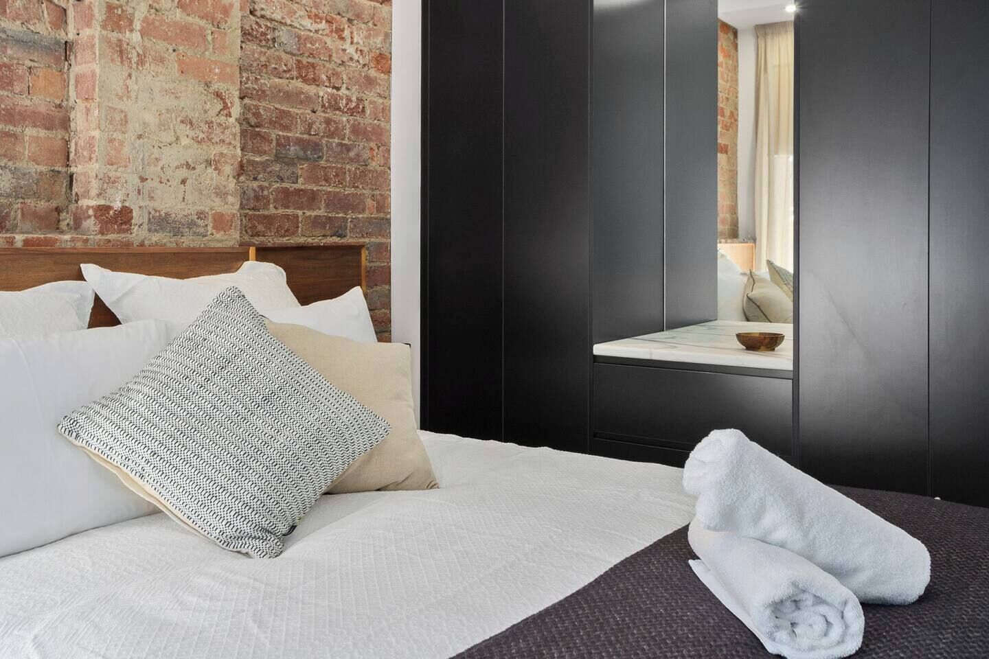 Une chambre d’hôtel avec un mur en briques, un lit avec du linge de maison blanc, une armoire en bois sombre et une petite table de chevet avec un bol.