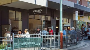 Ett utomhusområde med bord och stolar, en butiksfasad med skylten ”VILLINO COFFEE ROASTERS” och en flagga med texten ”City of Hobart”.