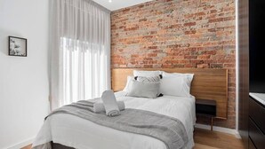 A modern bedroom with a brick wall, a wooden headboard, a bed with white bedding, and a large window with sheer curtains. - Sanctuary at Saddlers Where Rustic Meets Refined (Hobart)