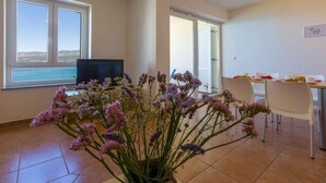 Ein modernes Wohnzimmer mit großem Fenster, einem Flachbildfernseher, einem Essbereich mit Tisch für zwei Personen und einem Blumenstrauß.