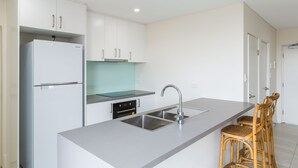 A modern kitchen with white cabinets, a stainless steel sink, and a built-in oven. There are two wooden bar stools and a small island with a glass top.