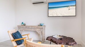 A room with a beach view on the TV, a wicker chair, a small table with a glass of wine, and a fireplace.