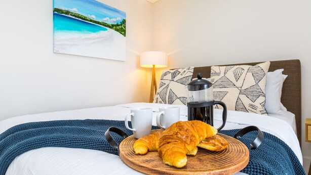 A neatly made bed with a blue blanket, a wooden tray with croissants and two white mugs, a coffee maker, and a painting of a beach on the wall. - Ishtar Apartment 8 by Book Jervis Bay - Luxury Studio (Huskisson)