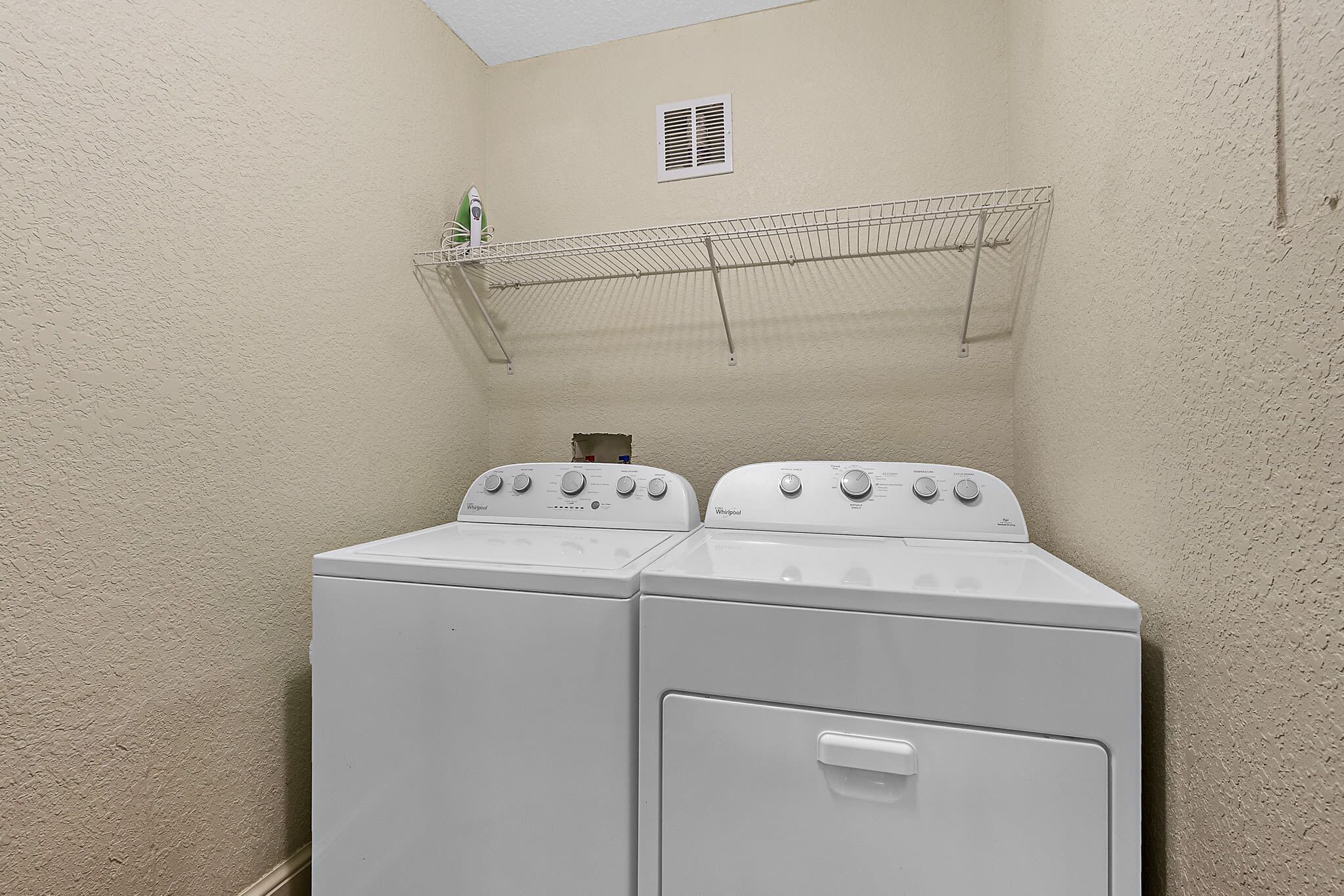 A laundry area with two white Whirlpool washers and dryers, a metal drying rack, and a vent on the wall.