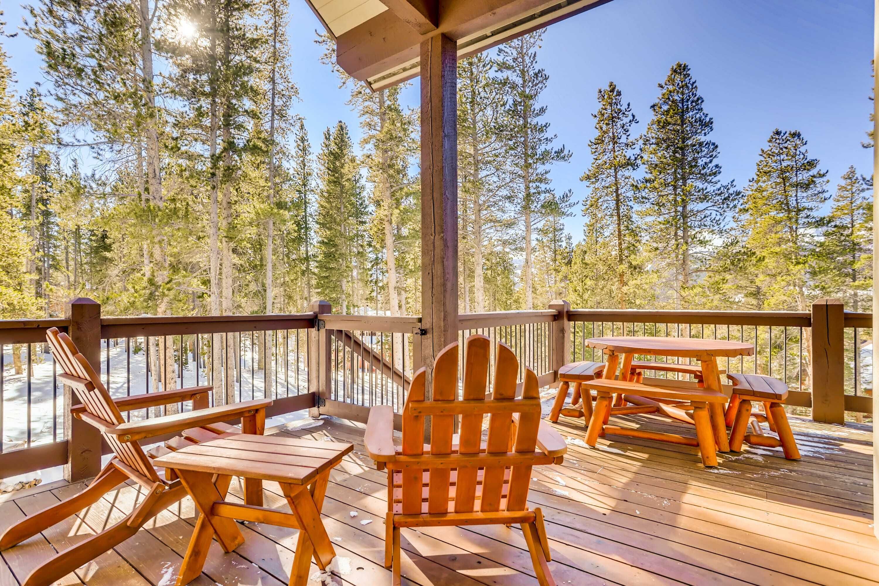 A wooden deck with Adirondack chairs and picnic tables, surrounded by tall pine trees.