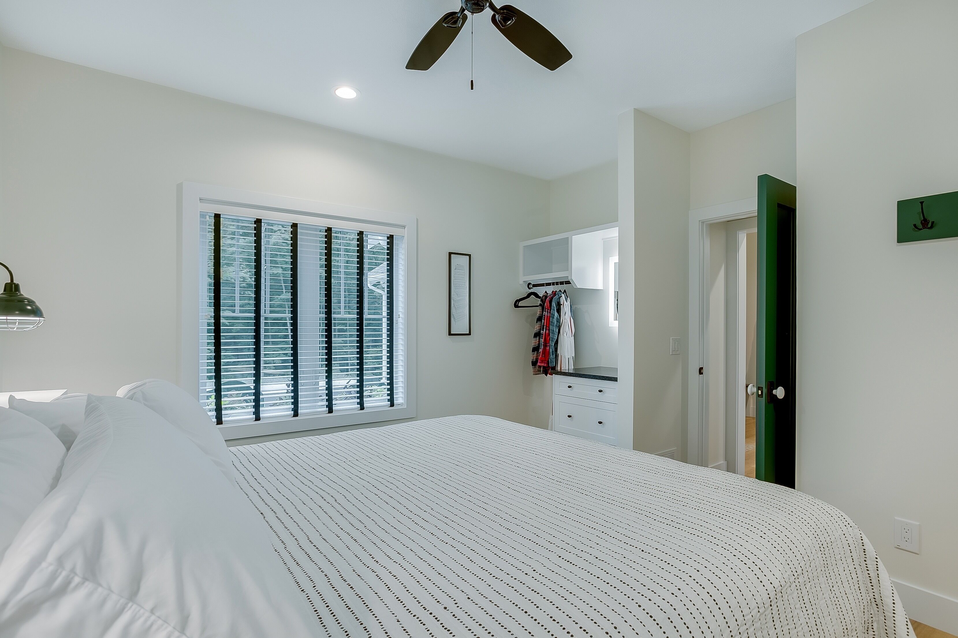 A bedroom with a bed, a ceiling fan, a window with blinds, a mirror, and a door.