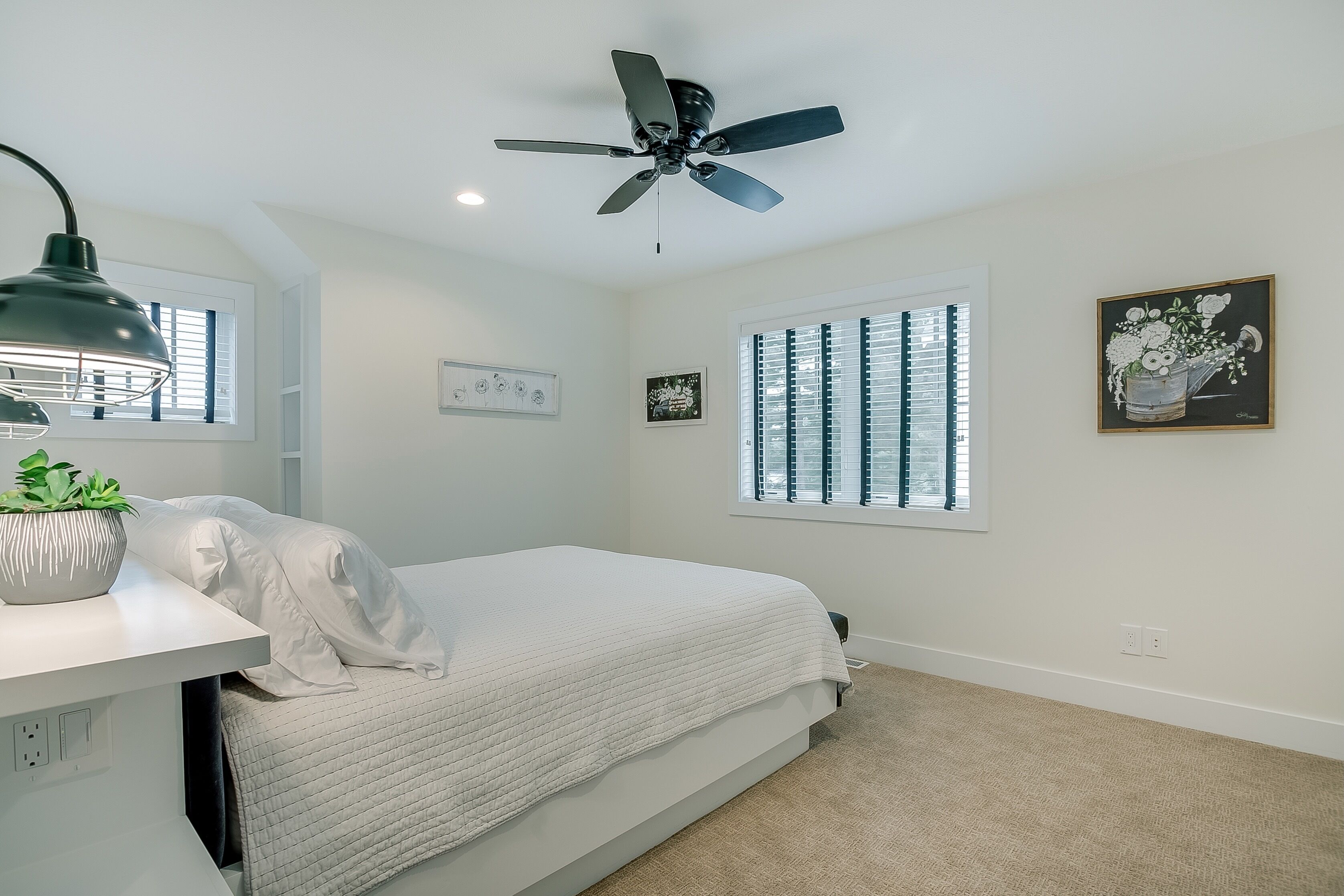 A bedroom with a bed, a ceiling fan, a desk with a lamp, and a window with blinds.