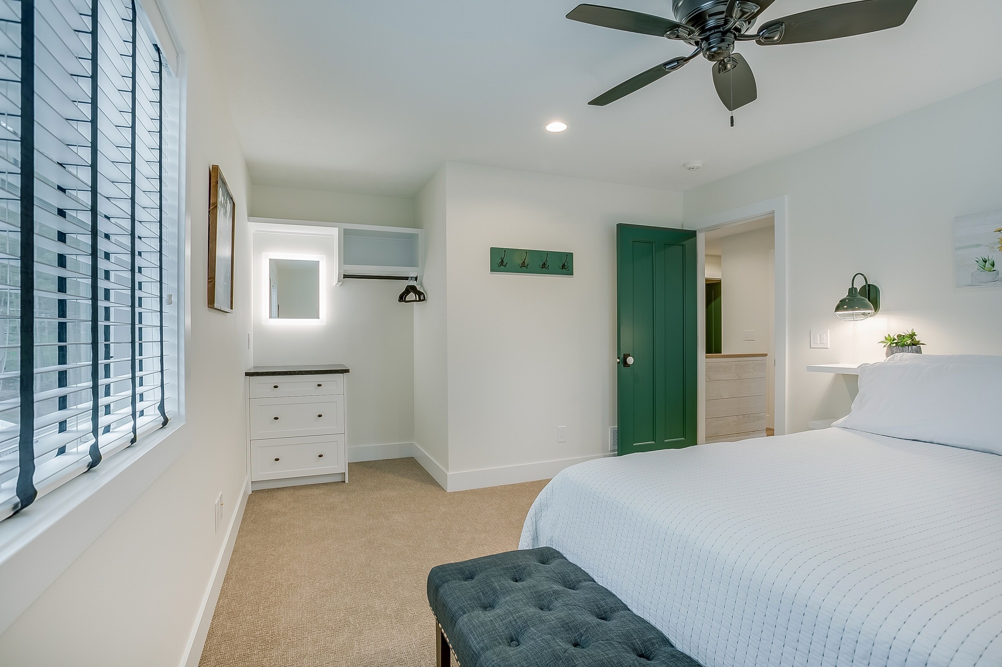 Un dormitorio con cama, ventilador de techo, cómoda, espejo, una puerta verde y una ventana con persianas.