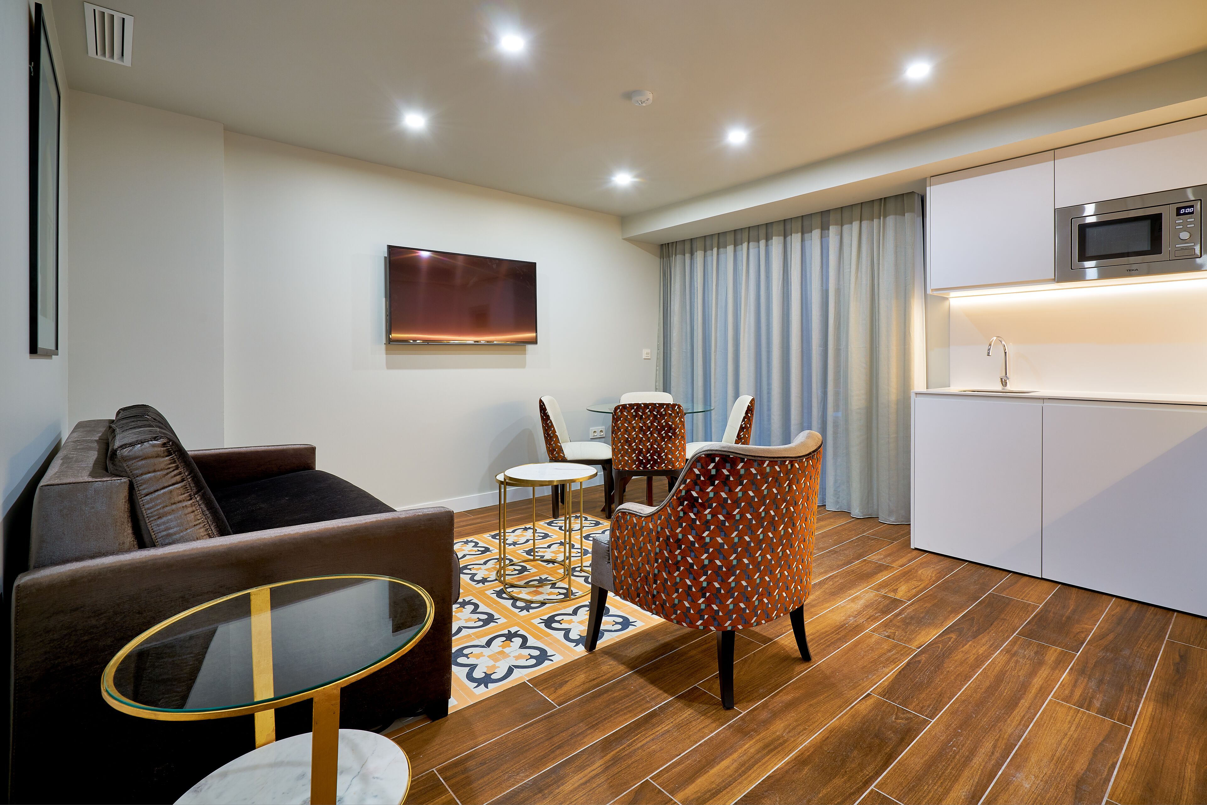 A modern living room with a sofa, a coffee table, patterned chairs, a flat-screen TV, and a kitchen area with a microwave.