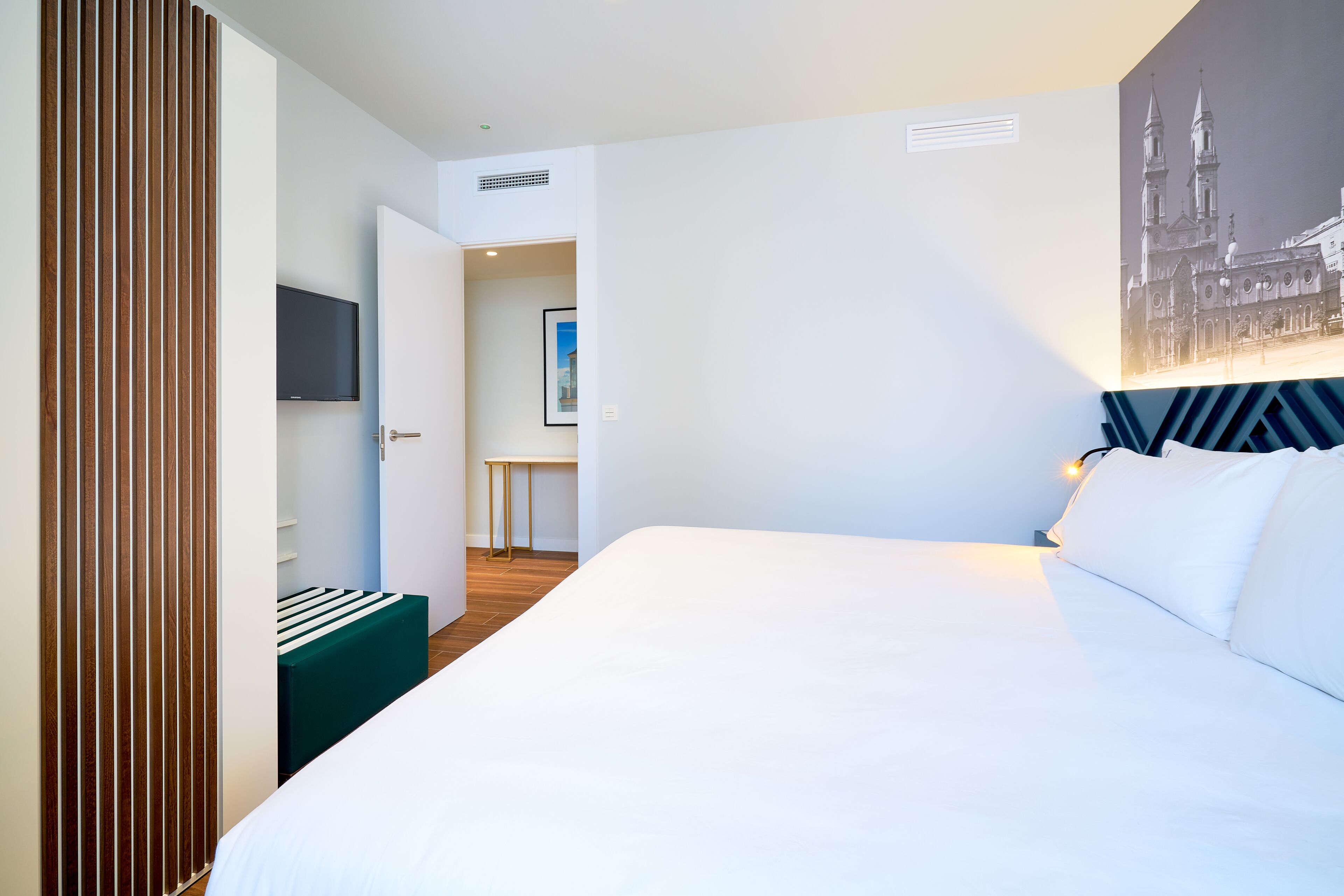 A modern hotel room with a large bed, a wooden slat partition, a flat-screen TV, and a framed picture on the wall.
