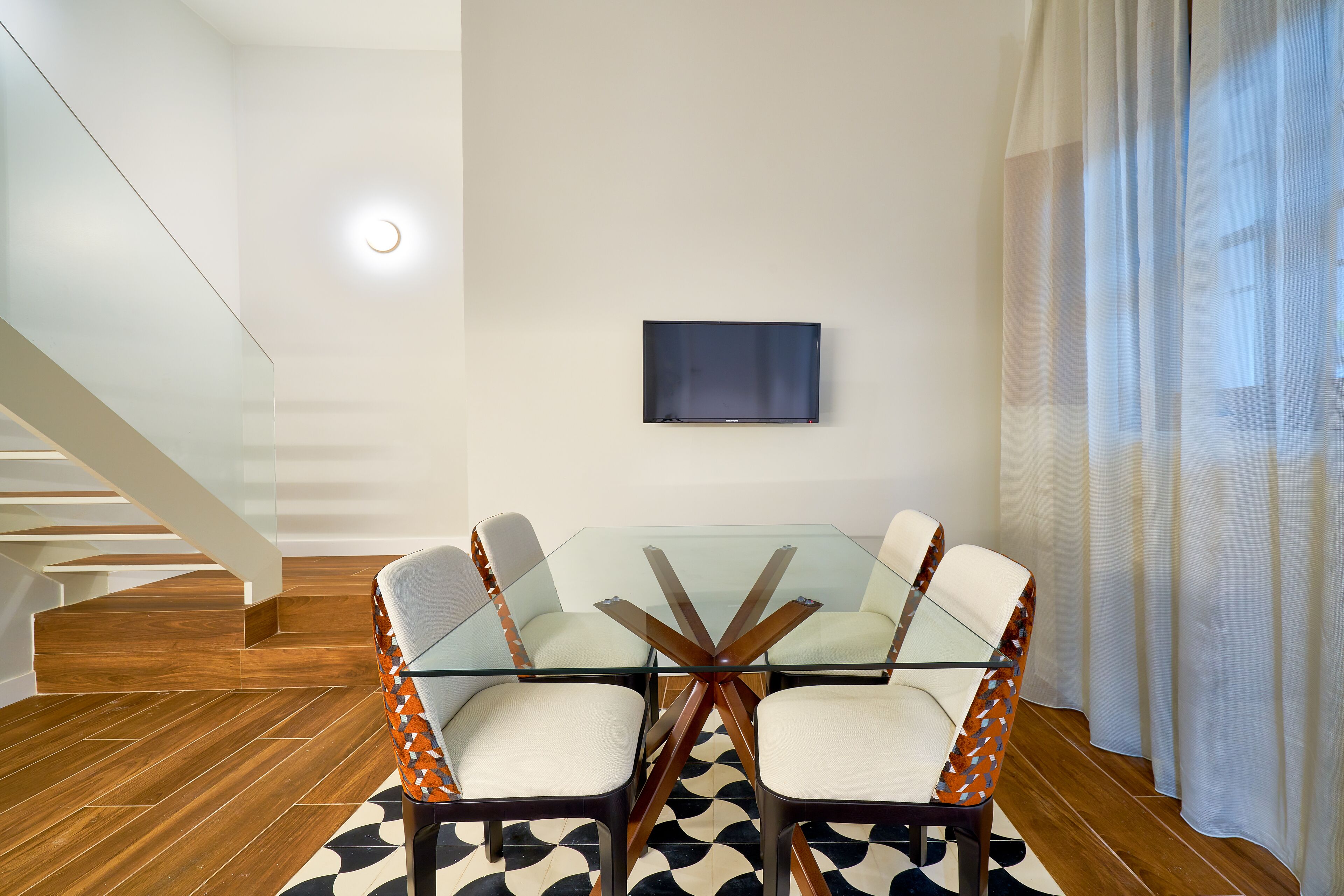 A modern dining area with a glass table and chairs, a flat-screen TV on the wall, and a staircase in the background.