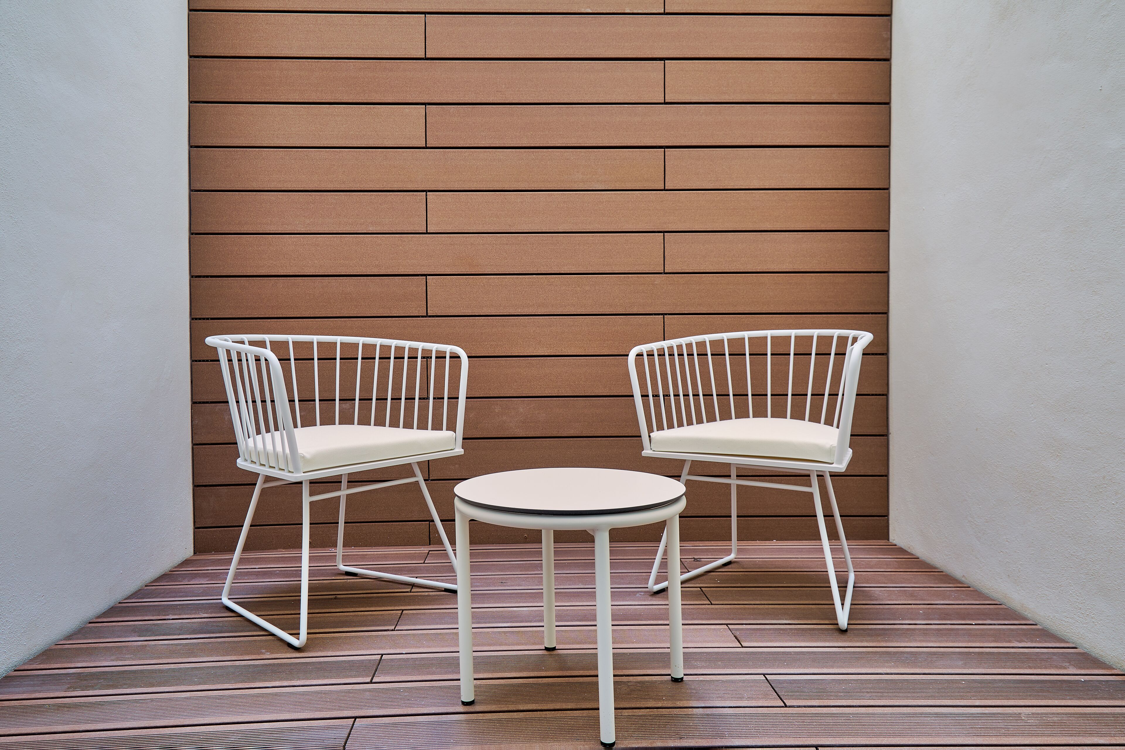 A small outdoor seating area with two white chairs and a round table, set against a wooden wall and wooden flooring.