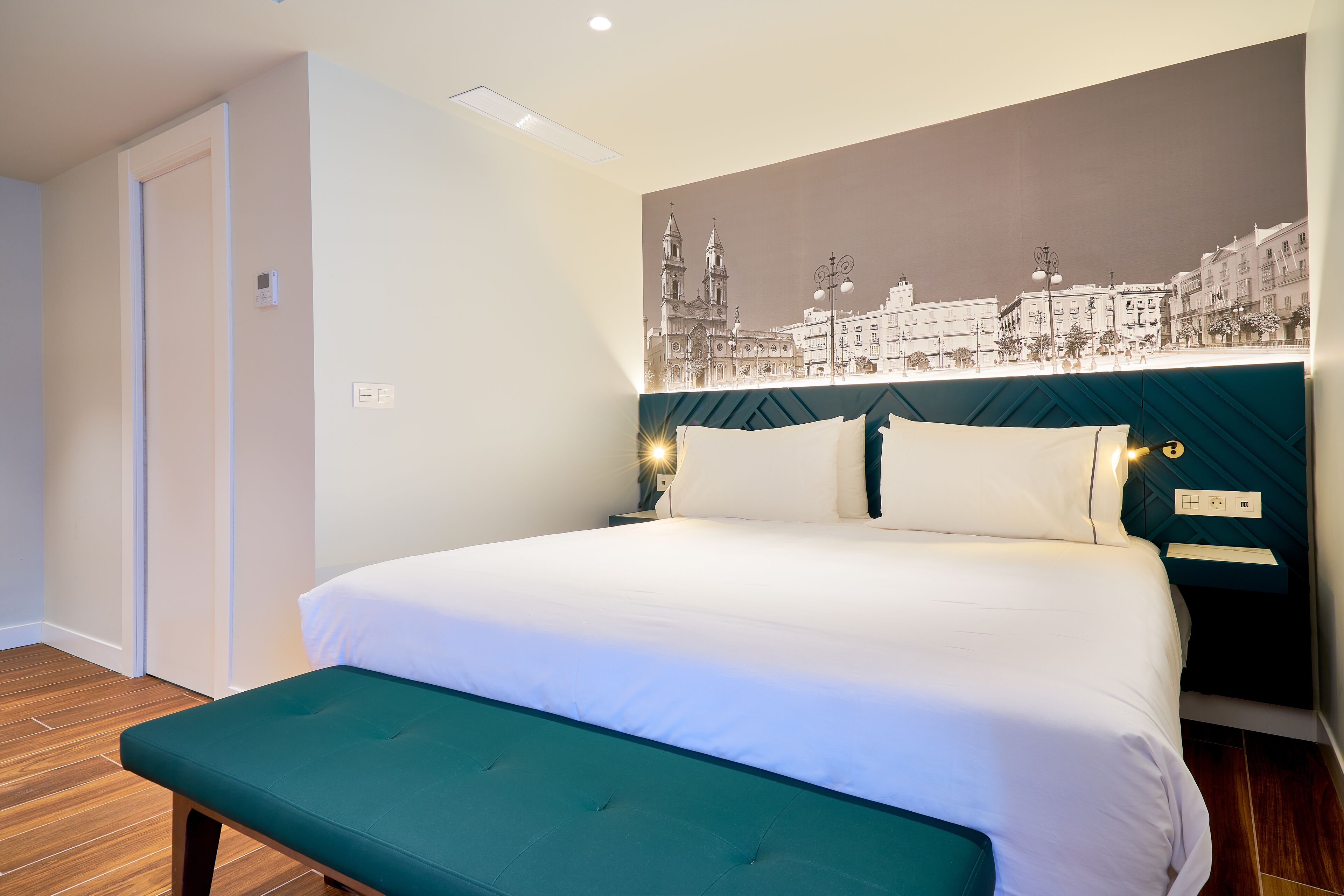 A neatly made bed with white linens, a teal bench, and a wall-mounted nightstand with a lamp.