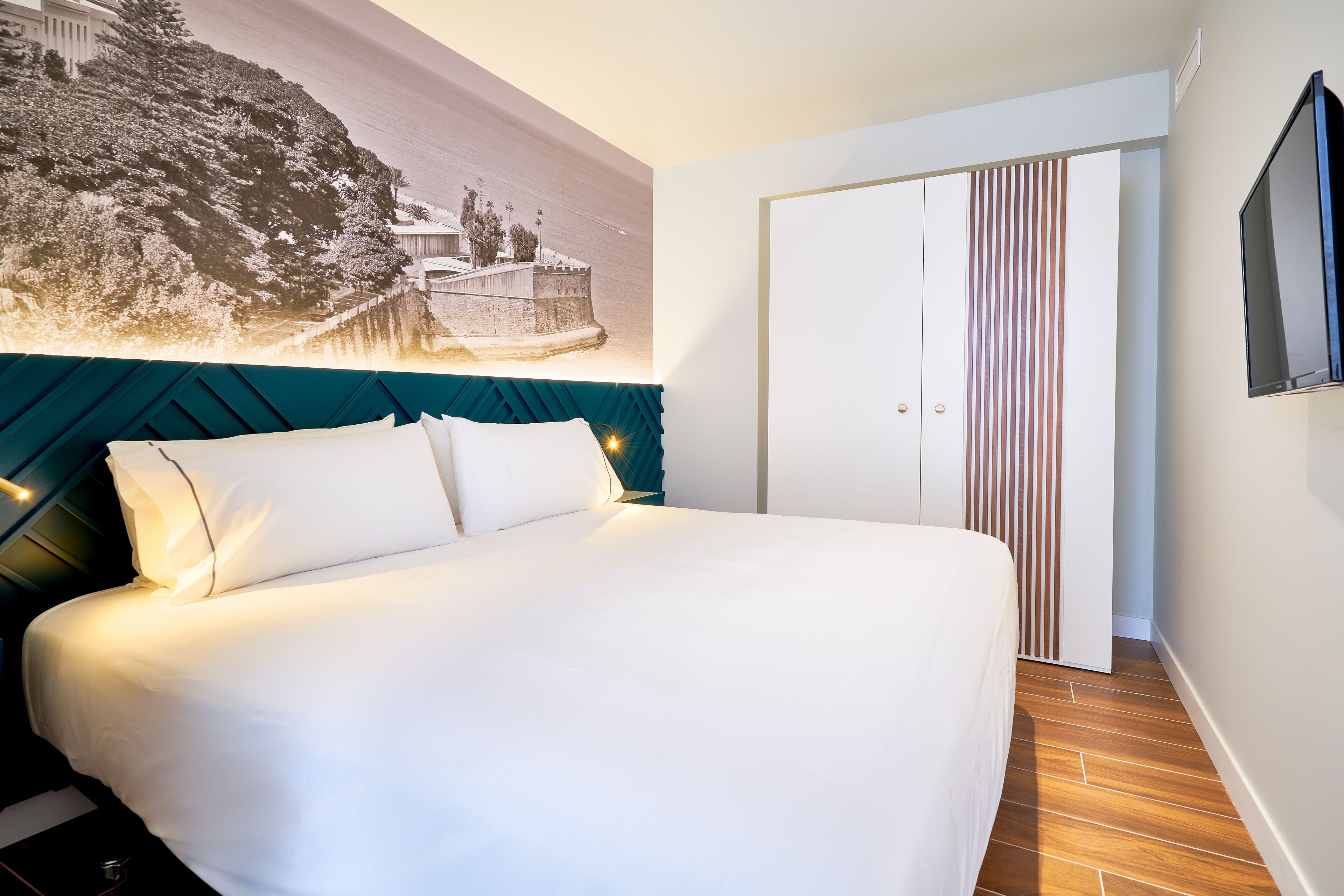 Een net opgemaakt bed met witte lakens, een blauwgroen hoofdbord en een groot muurschildering van een besneeerd landschap.