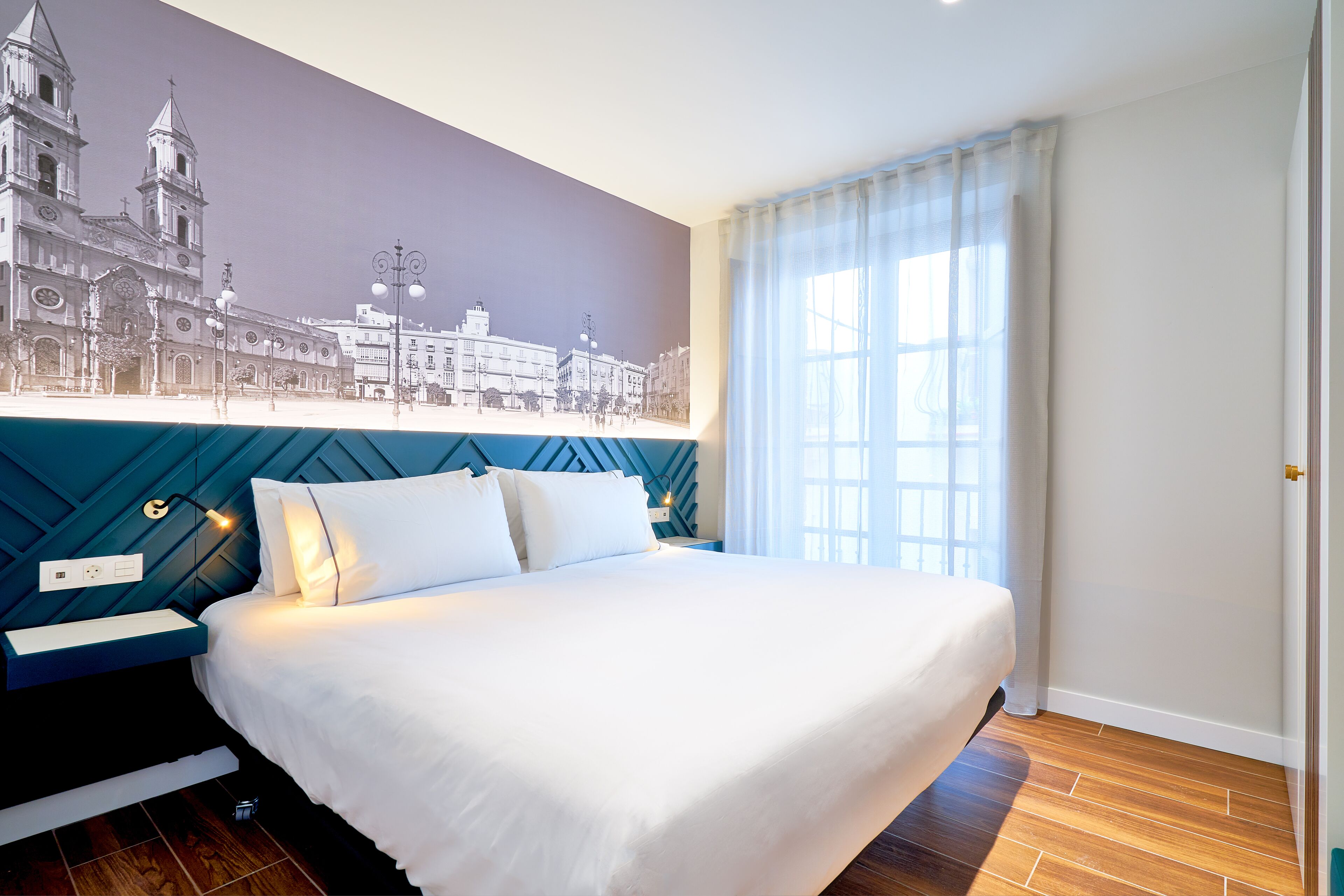 A modern hotel room with a large bed, a headboard with a cityscape mural, a bedside table with a lamp, and a window with curtains.