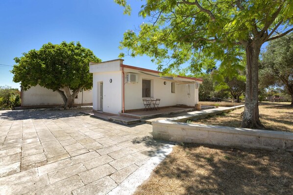 A small, single-story house with a tiled patio, a small table and chairs outside, and a tree in the foreground.