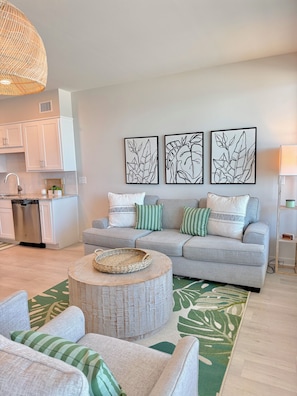 A living room with a grey sofa, a round coffee table, and a kitchen area with white cabinets and a dishwasher.