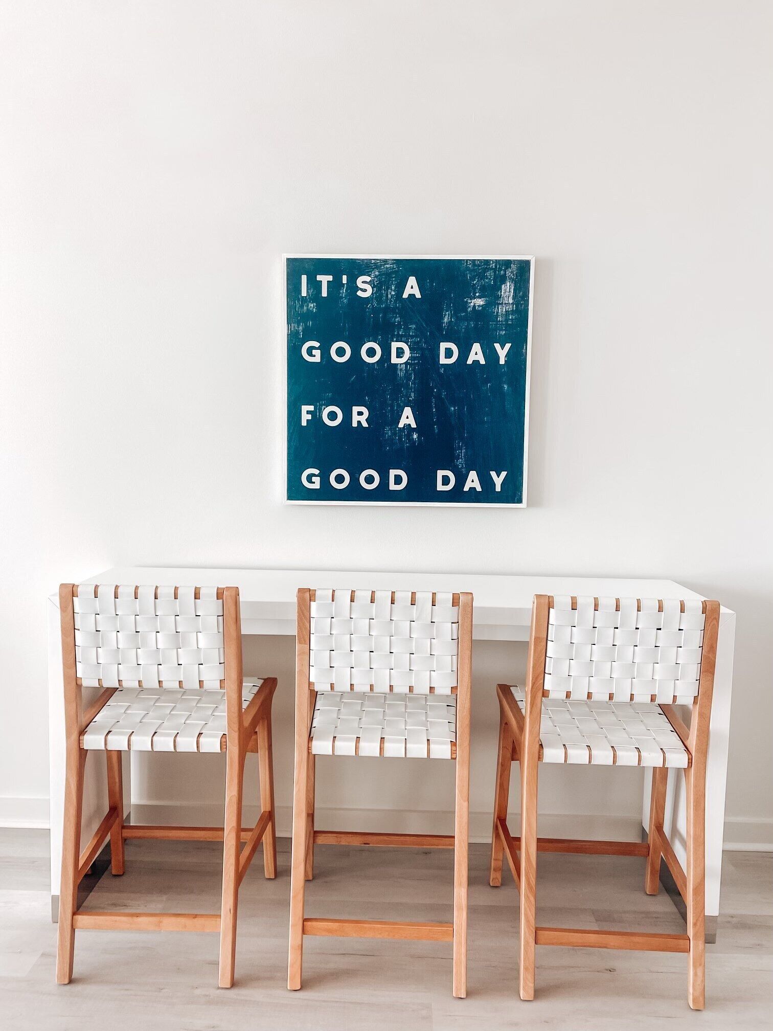 Drei weiße, gewebte Stühle mit Holzgestell vor einer weißen Wand. Darauf eine blaue Schilder mit der Aufschrift 'IT'S A GOOD DAY FOR A GOOD DAY'.