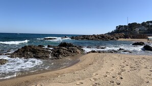 Une plage avec des affleurements rocheux, un ciel bleu dégagé et un bâtiment visible au loin.