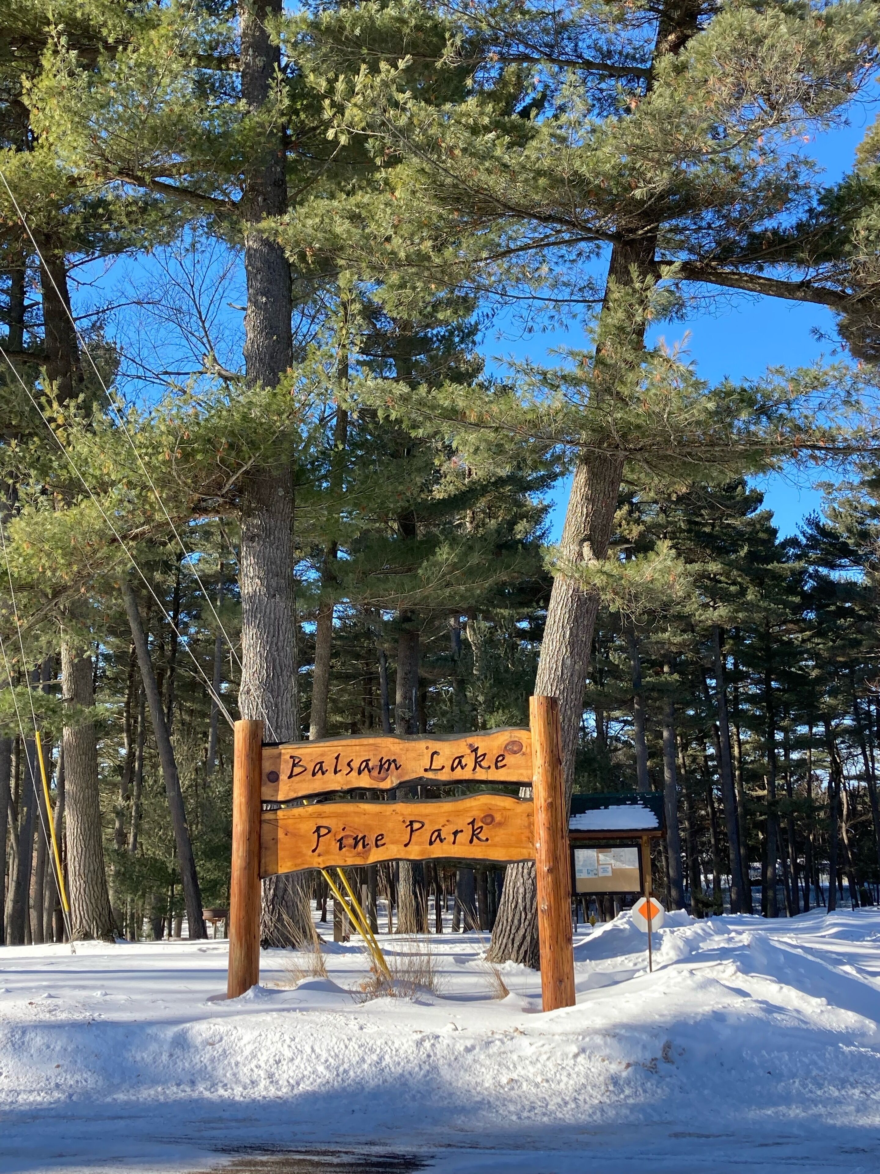 A wooden sign with 'Balsam Lake Pine Park' written on it, surrounded by snow and trees.