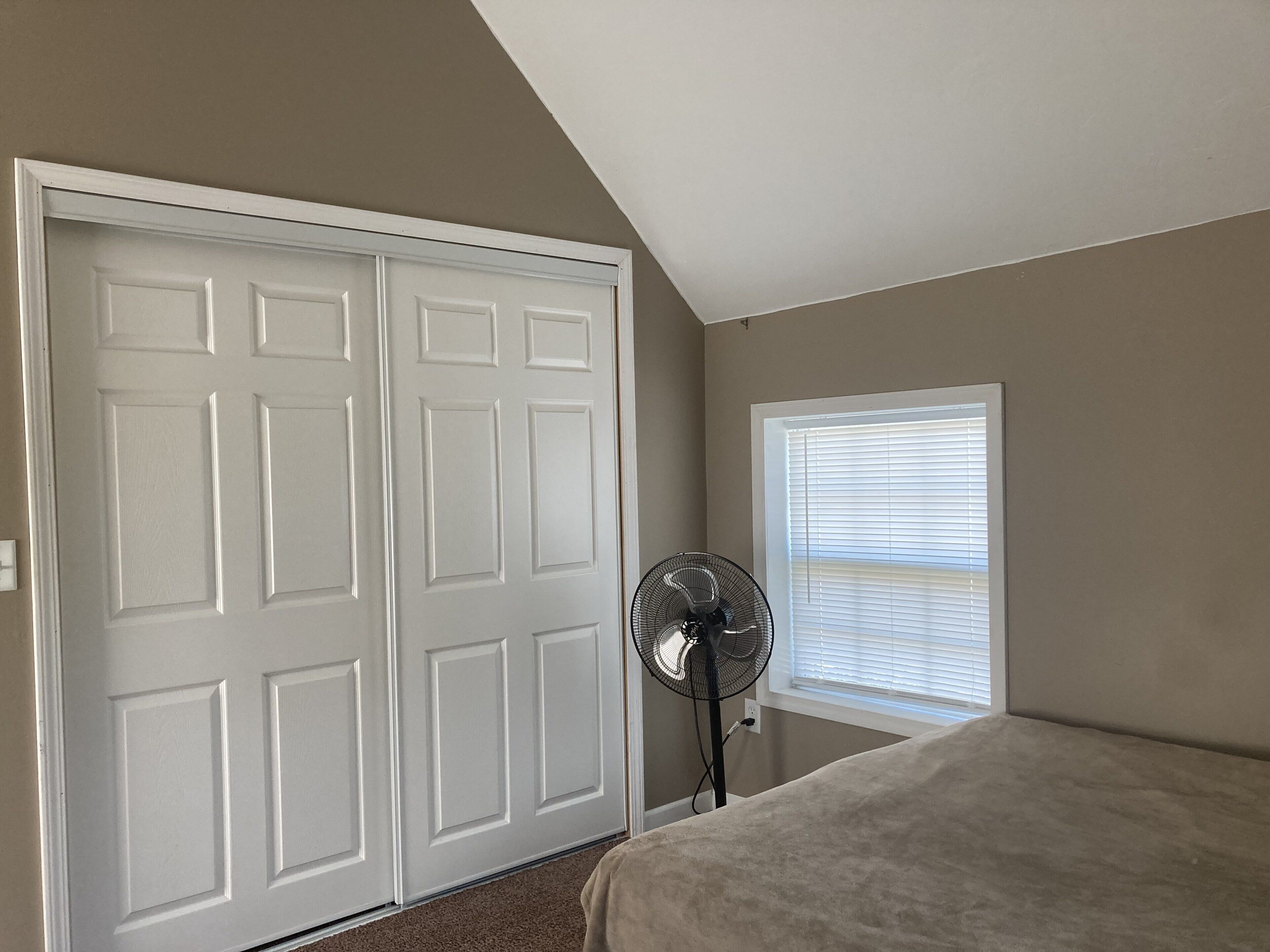 Un dormitorio con una puerta blanca, una ventana con persianas y un ventilador de pie.