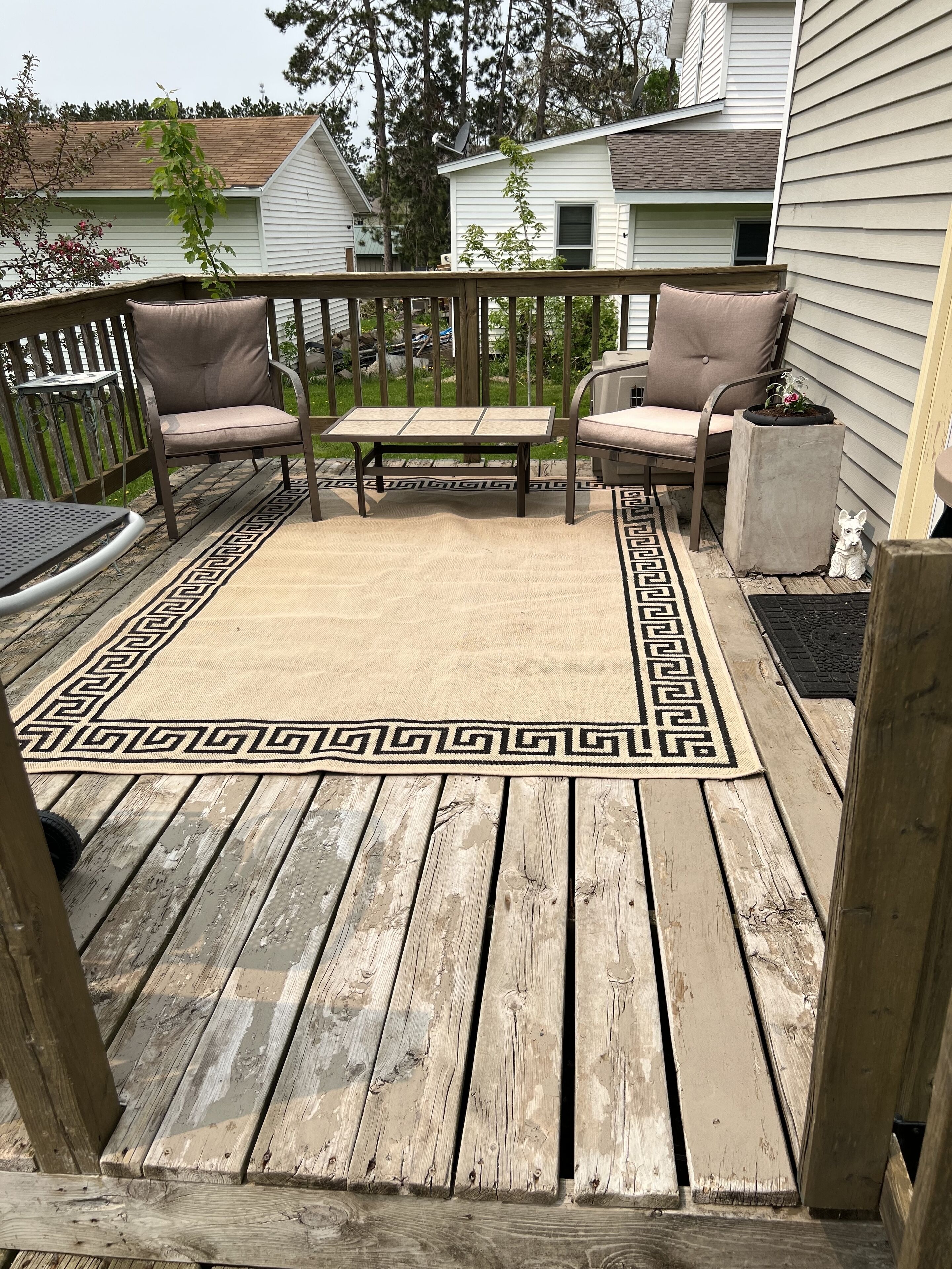 A wooden deck with a patterned rug, two chairs, a small table, and a potted plant.