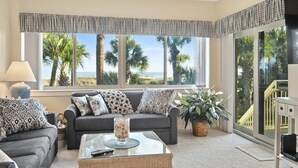 A living room with a sofa, a coffee table, and a large window offering a view of palm trees and the beach. - 403 Captains Walk - Palmetto Dunes 1st Floor Oceanfront - Hilton Head Rentals (Hilton Head Island)