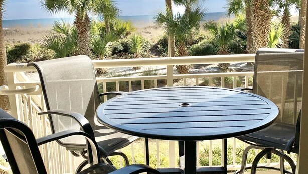 A deck with a round table and chairs overlooking a beach with palm trees. - 403 Captains Walk - Palmetto Dunes 1st Floor Oceanfront - Hilton Head Rentals (Hilton Head Island)