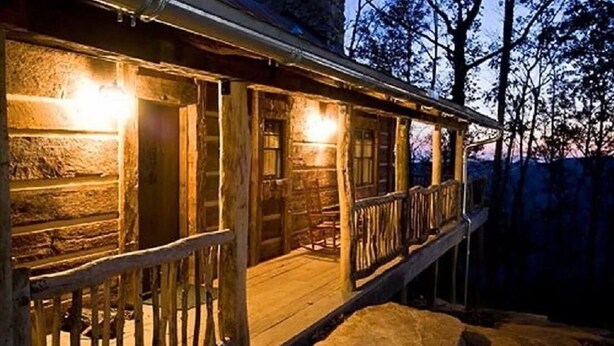 Une cabane en bois rustique, dotée d’un porche, de fondations en pierre et offrant une vue sur la forêt environnante.