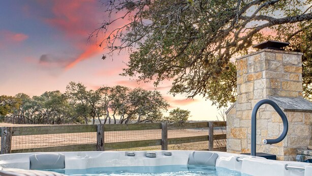 Un jacuzzi con chimenea de piedra, rodeado por una valla de madera y árboles, con un atardecer al fondo.