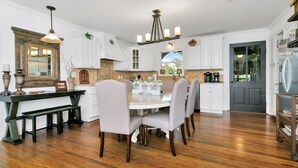 Una cocina con muebles blancos, una mesa de comedor con sillas, una araña y vistas al exterior.