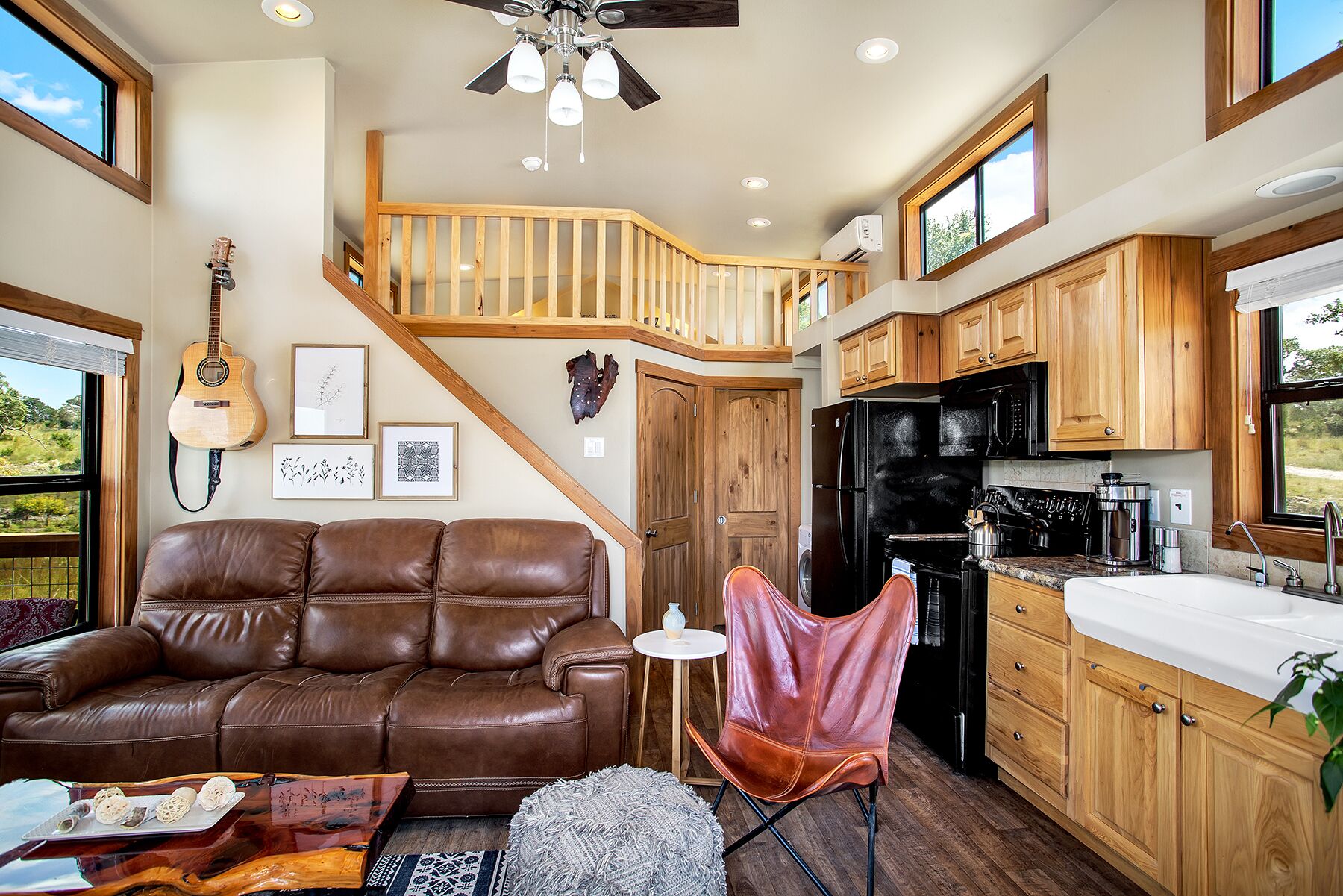 A spacious living room with a brown leather sofa, a small round rug, and a wooden coffee table. A red leather chair is positioned near the kitchen area, which features wooden cabinets, a black refrigerator, and a countertop with a sink.