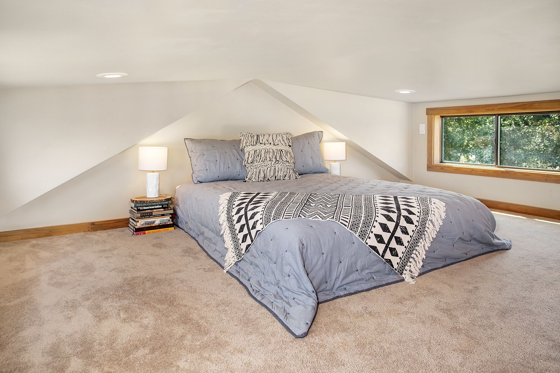 Een slaapkamer met een schuine plafond, een bed met een grijze spreidek en kussens, een nachtkastje met een lamp, en een raam met uitzicht op bomen.
