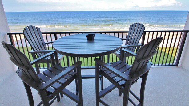 A round table with four Adirondack chairs on a balcony overlooking the ocean.