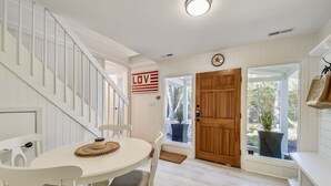 Un coin repas avec une table et des chaises blanches, une porte en bois et une vue sur l’extérieur.