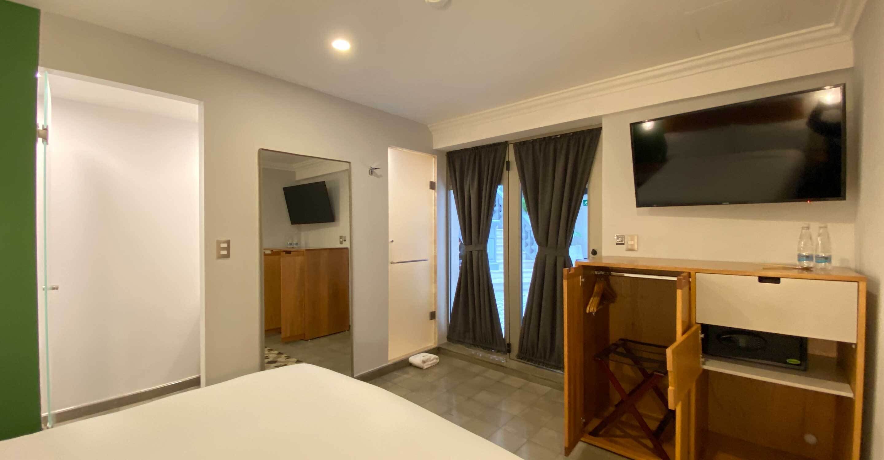 Habitación de hotel con una cama, un televisor montado en la pared, un tocador de madera y una puerta que da a un balcón.