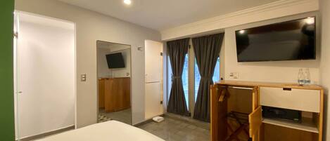 Habitación de hotel con una cama, un televisor montado en la pared, un tocador de madera y una puerta que da a un balcón.