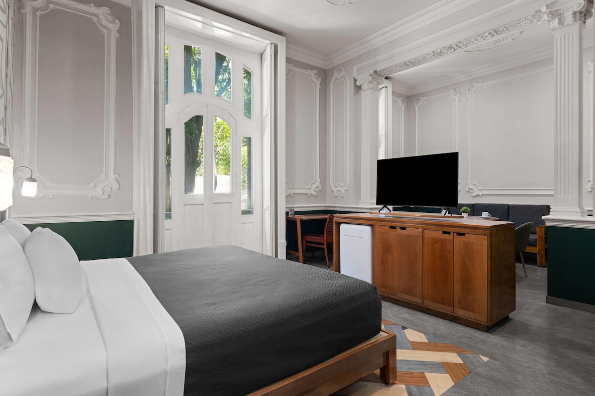 Moderni hotellihuone, jossa on sänky, televisio, pieni keittonurkkaus ja näkymä vihreälle alueelle.