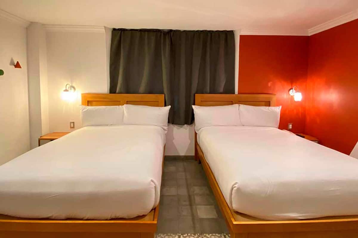Een hotelkamer met twee eenpersoonsbedden, een raam met donkere gordijnen, een rode accentmuur en bedlampen.