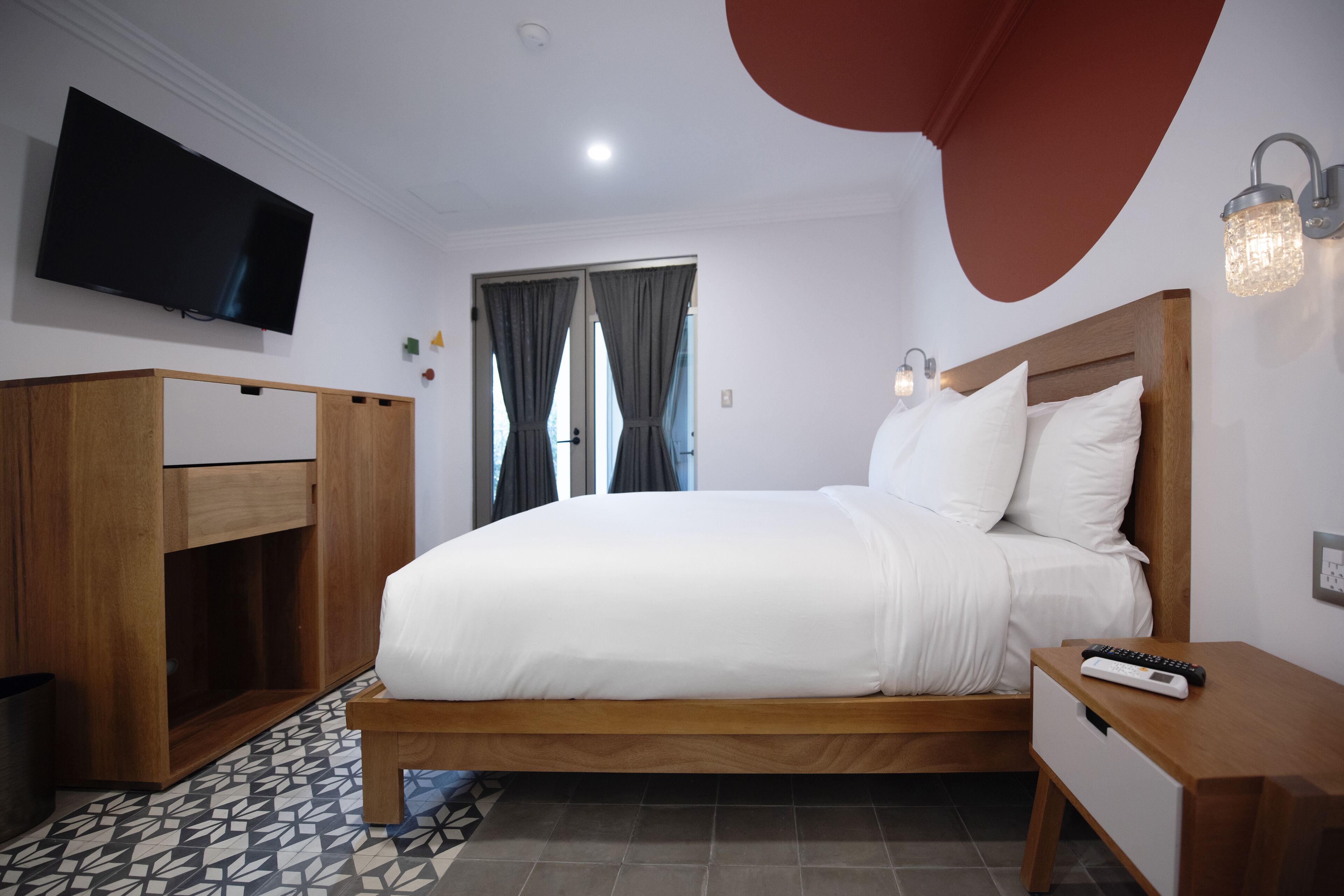 A hotel room with a bed, a TV mounted on the wall, a wooden wardrobe, a bedside table, and a window with curtains.