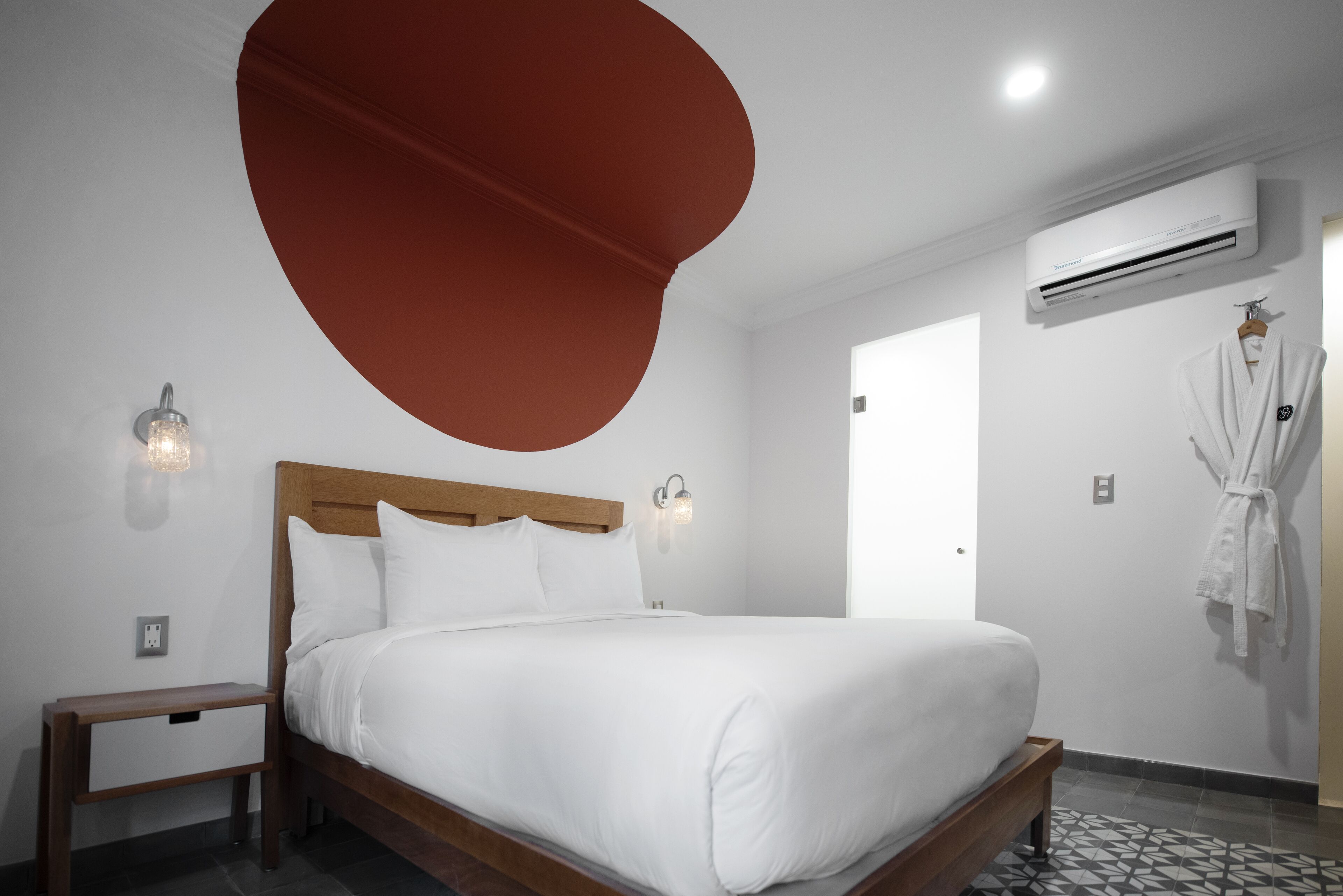 A neatly made bed in a hotel room with a wooden headboard, a nightstand, an air conditioning unit, and a robe hanging on the wall.