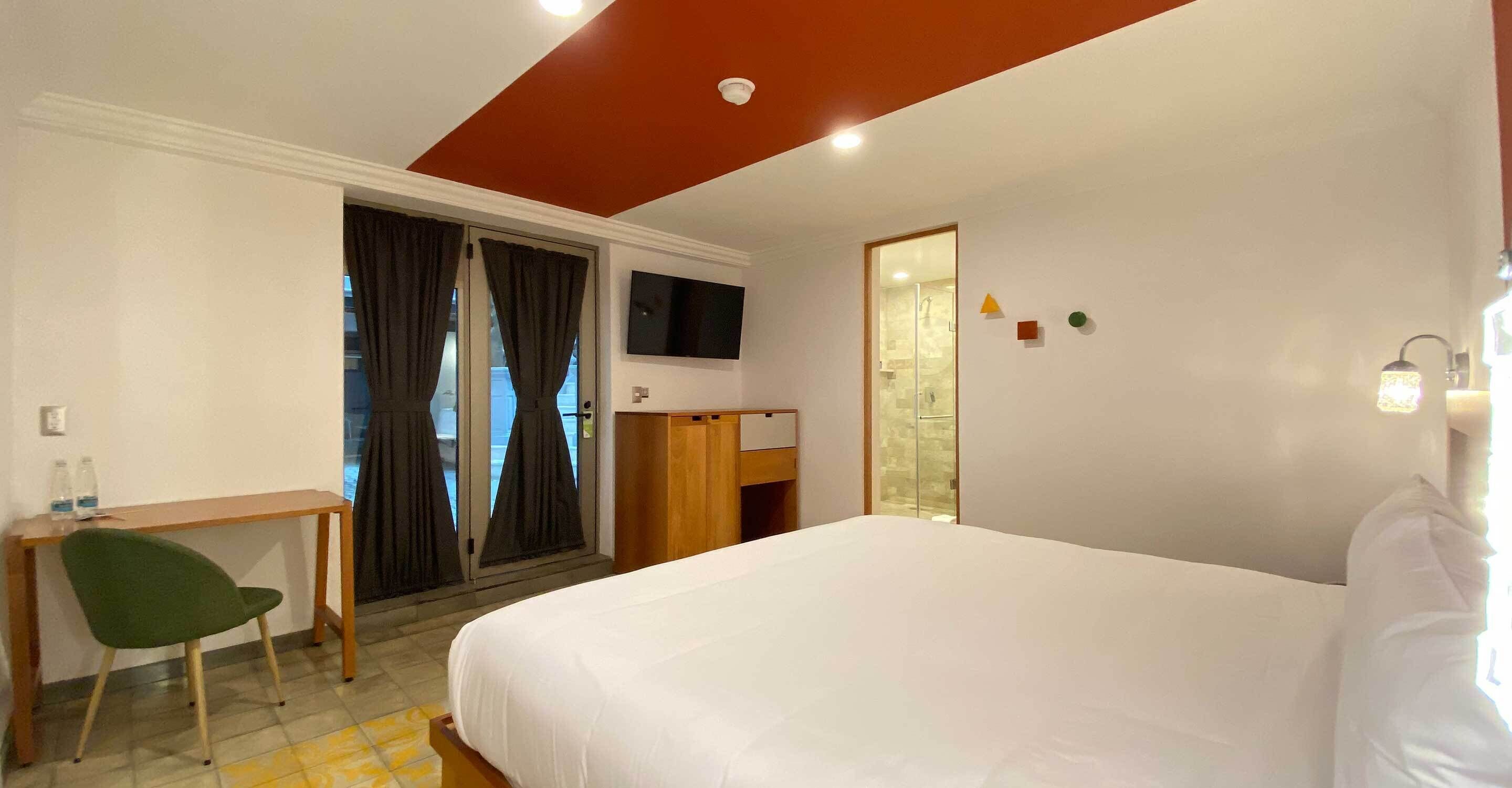 A hotel room with a bed, a desk, a chair, a TV, and a bathroom visible through a glass door.