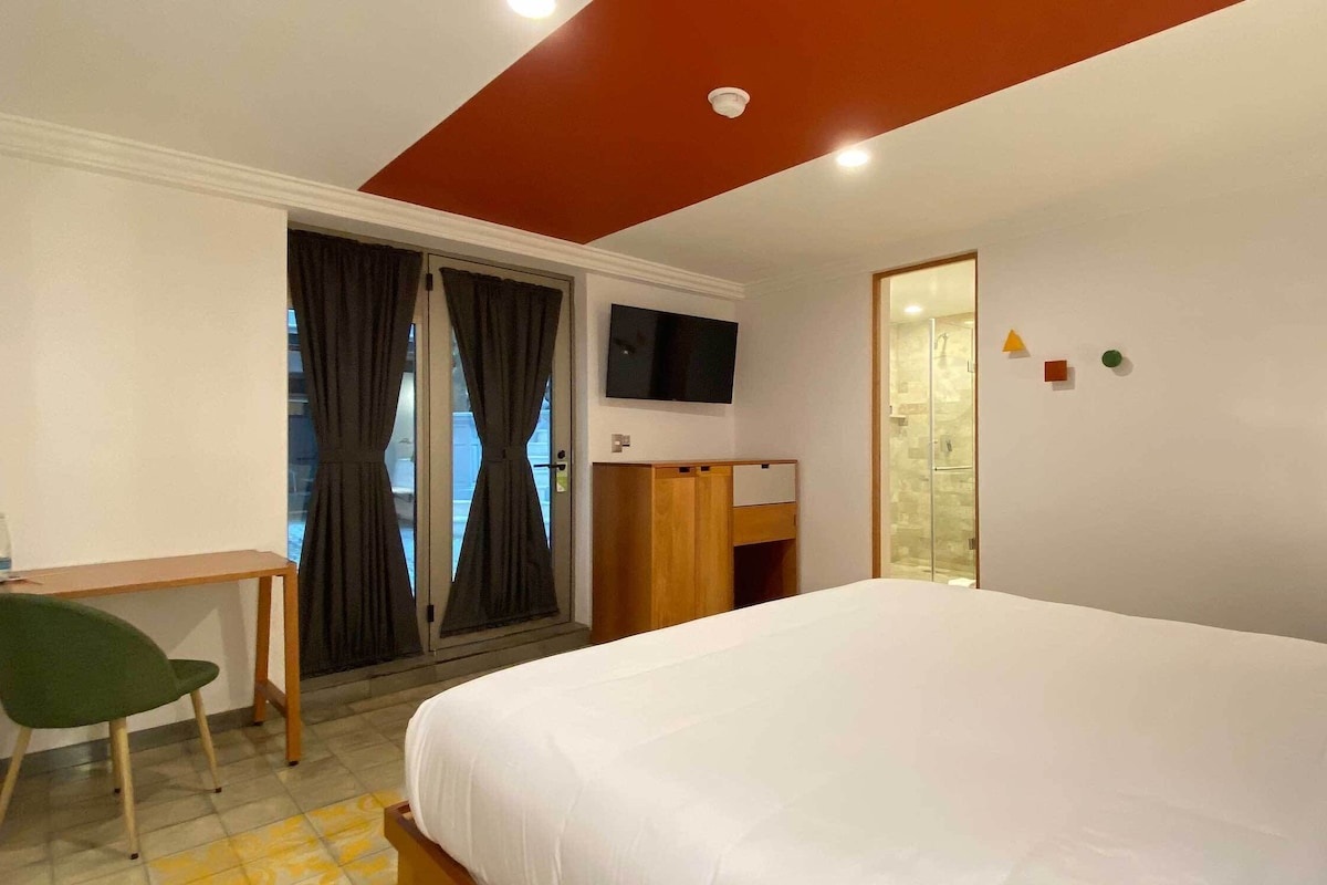 Habitación de hotel con cama, escritorio, silla, televisión y baño visible a través de una puerta de cristal.