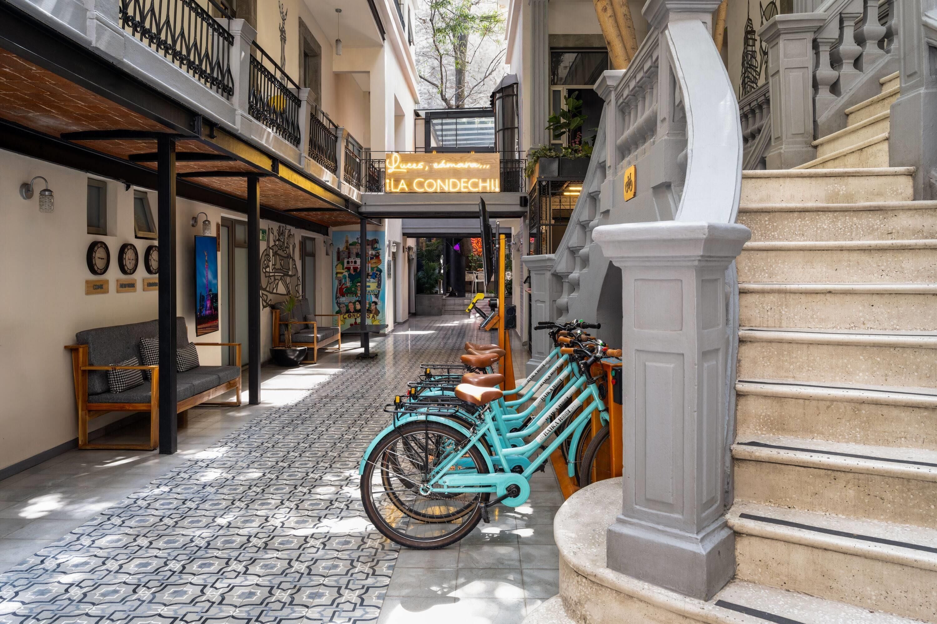 Une rue étroite bordée d’une rangée de vélos turquoise garés sur le trottoir, un escalier avec une balustrade blanche et un bâtiment orné d’une enseigne au néon sur laquelle on peut lire « LA CONDECHU ».