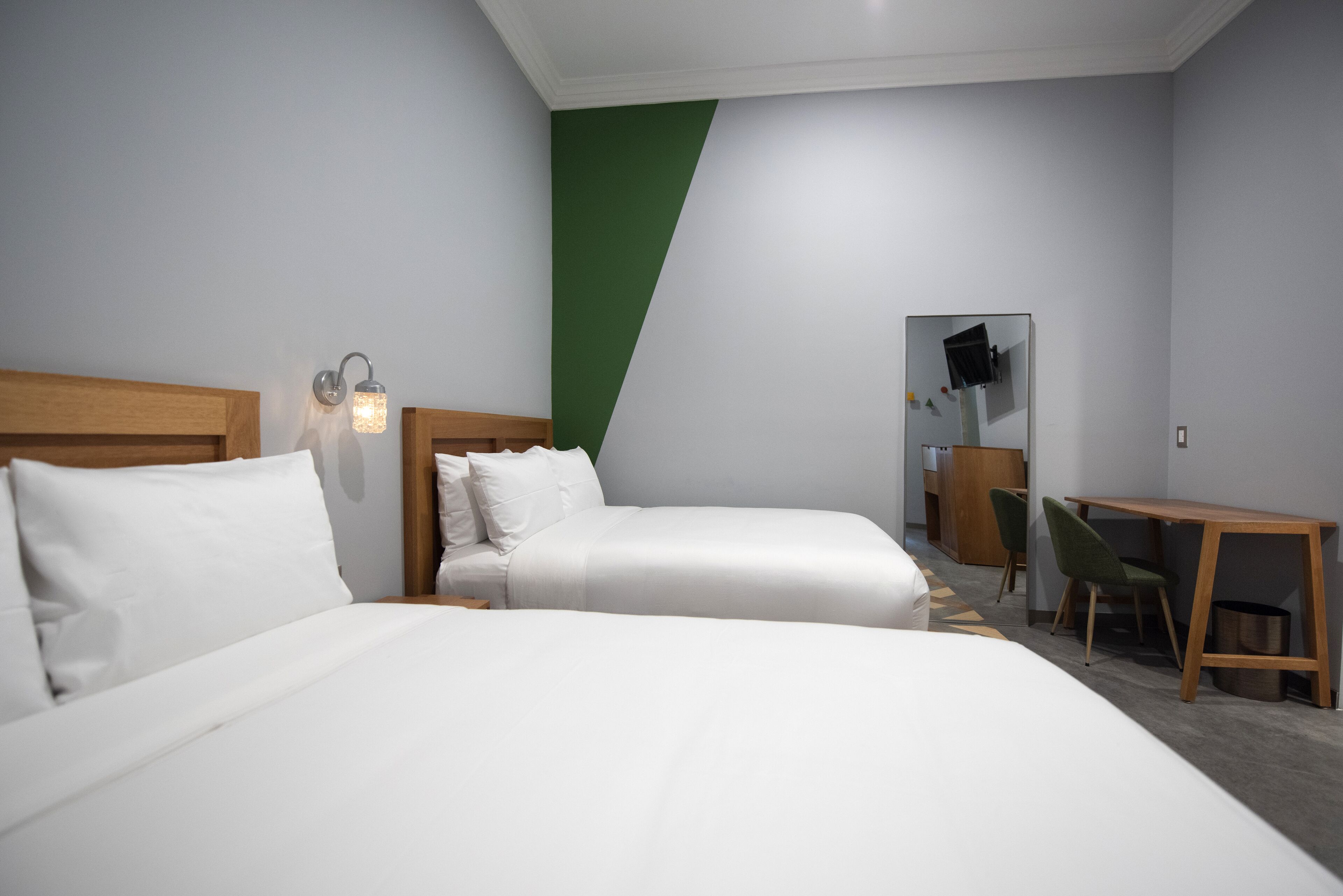 A hotel room with two beds, a desk, a chair, a mirror, and a green accent wall.
