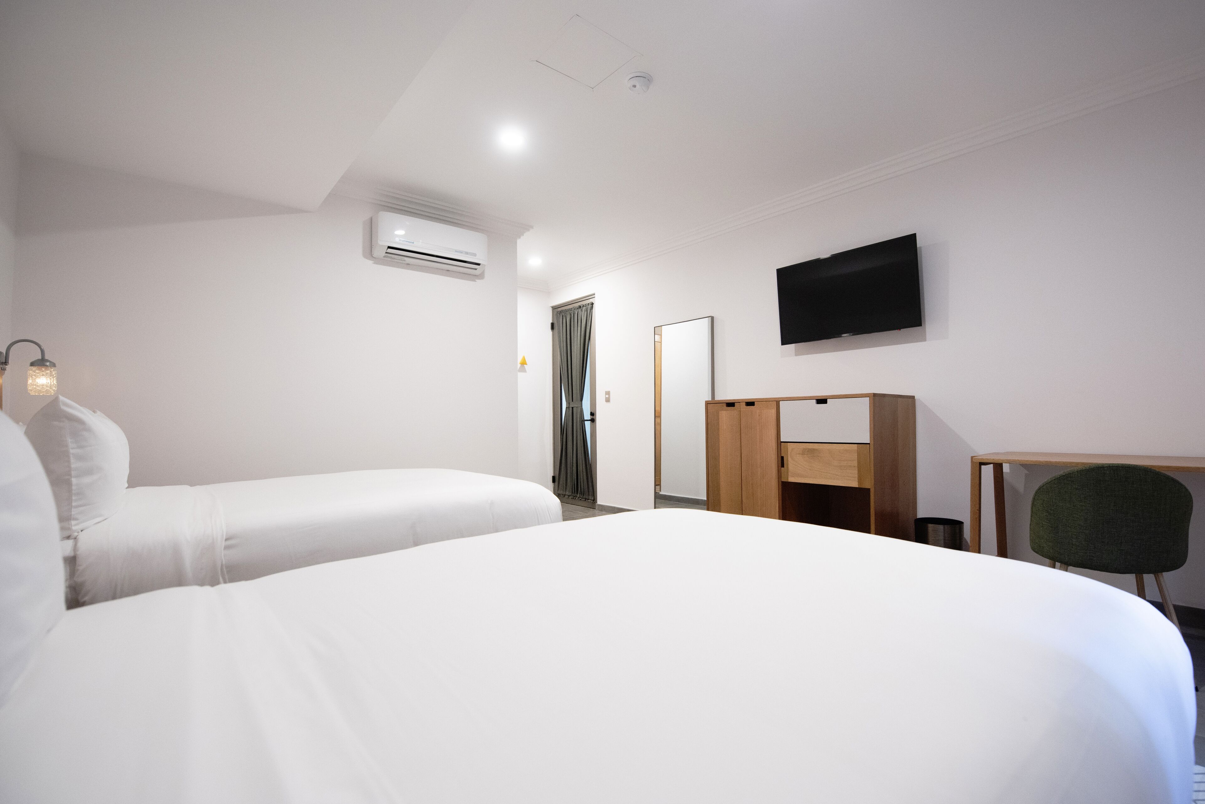 A hotel room with two single beds, a desk, a chair, a television, and an air conditioning unit.