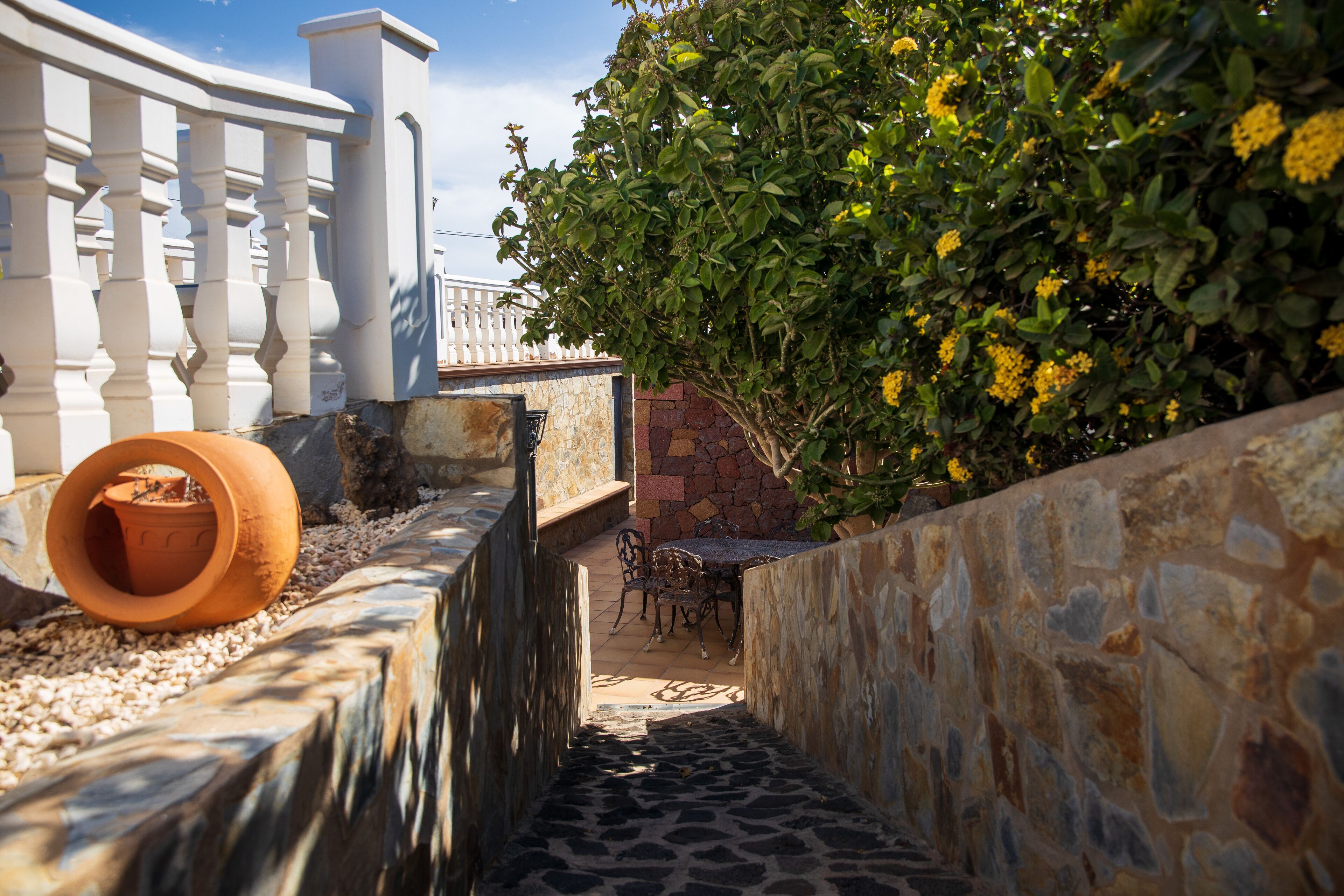 A narrow stone pathway leading to a patio area with a table and chairs, surrounded by a white railing and a stone wall.