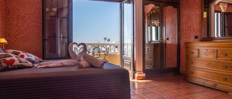 A bedroom with a large bed, a dresser, and a balcony with a view.